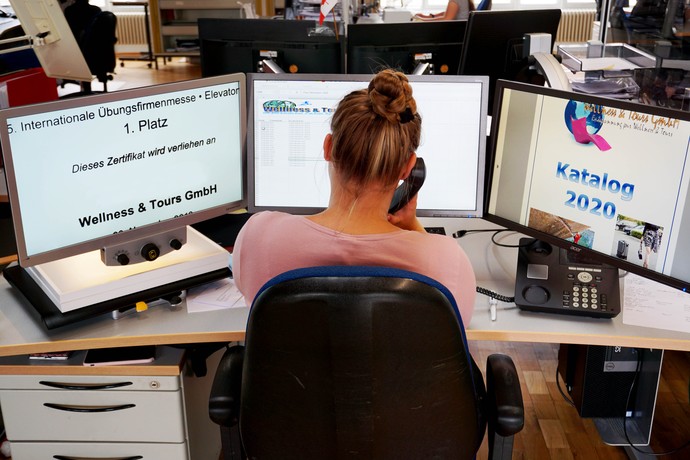 Eine Teilnehmerin sitzt an ihrem Schreibtisch mit 2 PCs und einem Bildschirmlesegerät zur Vergrößerung und telefoniert