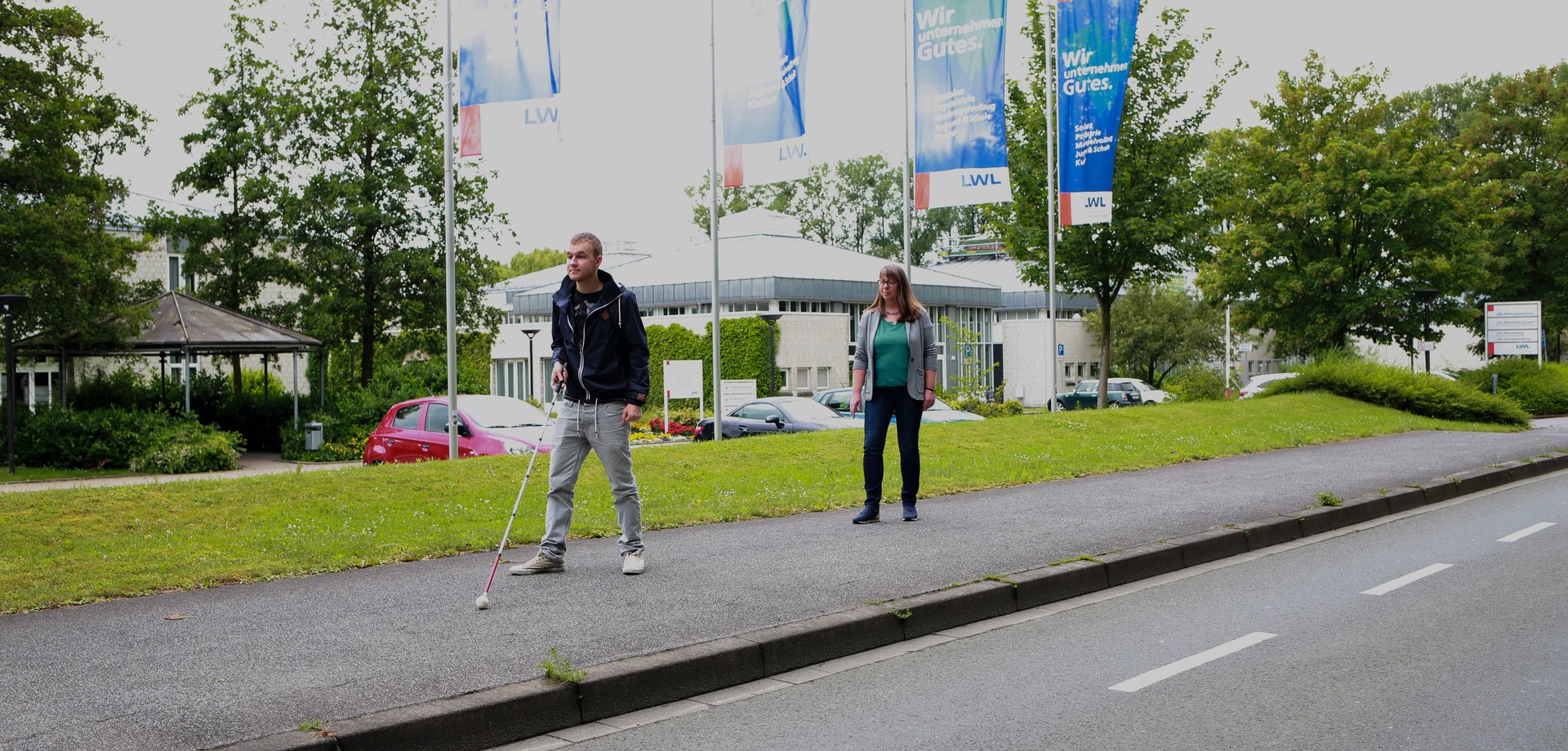 Ein Teilnehmer geht mit seinem Langstock auf dem Gehweg vor dem BBW entlang. Dahinter folgt seine Ausbilderin.
