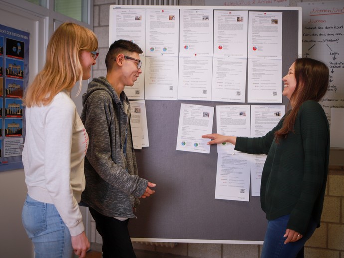 Eine Teilnehmerin und ein Teilnehmer stehen mit einer Lehrkraft vor einer Pinnwand