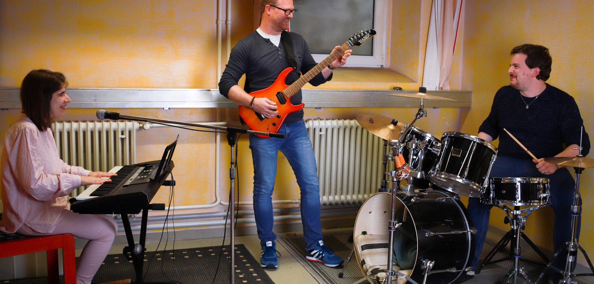 Eine Teilnehmerin sitzt am Keyboard, ein Teilnehmer spielt Gitarre und ein Teilnehmer sitzt am Schlagzeug im Proberaum