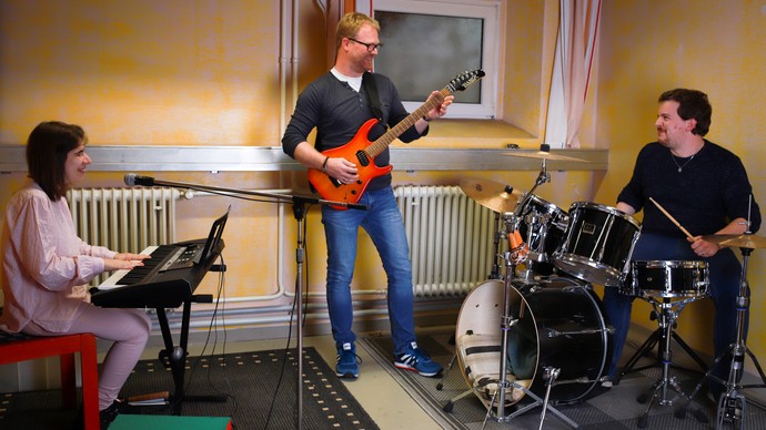 Eine Teilnehmerin sitzt am Keyboard, ein Teilnehmer spielt Gitarre und ein Teilnehmer sitzt am Schlagzeug im Proberaum