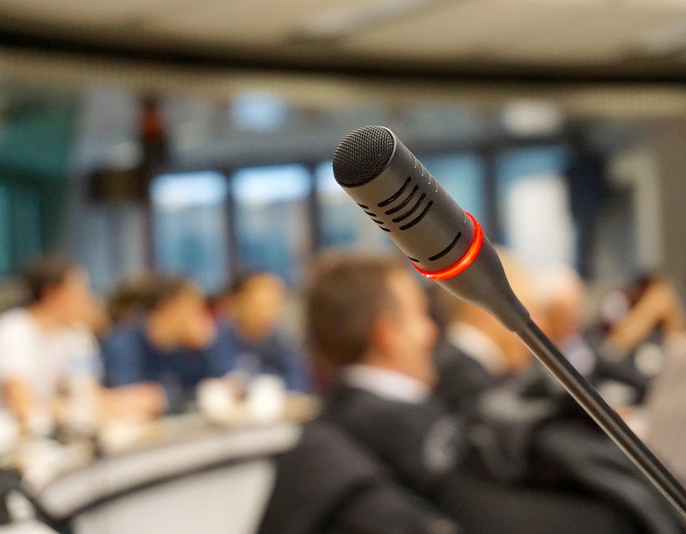 Im Vordergrund ein Mikrofon, im Hintergrund verschwommene Menschen