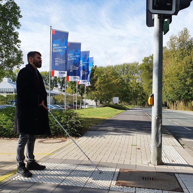 Ein Mann mit Langstock wartet an der Ampel vor dem BBW-Gebäude.