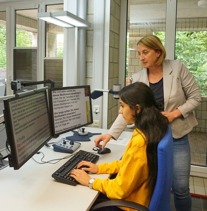 Die Facheberaterin hilft einer Teilnehmerin bei der Bildschirmarbeit. Der Arbeitsplatz ist ausgestattet mit Hilfsmitteln.